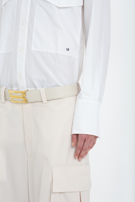 Close-up of a person wearing a white long-sleeved shirt, Victoria Beckham Relaxed Cargo Trouser In Bone with a yellow buckle belt and a large pocket on the side. The person’s right hand is visible near the pocket.