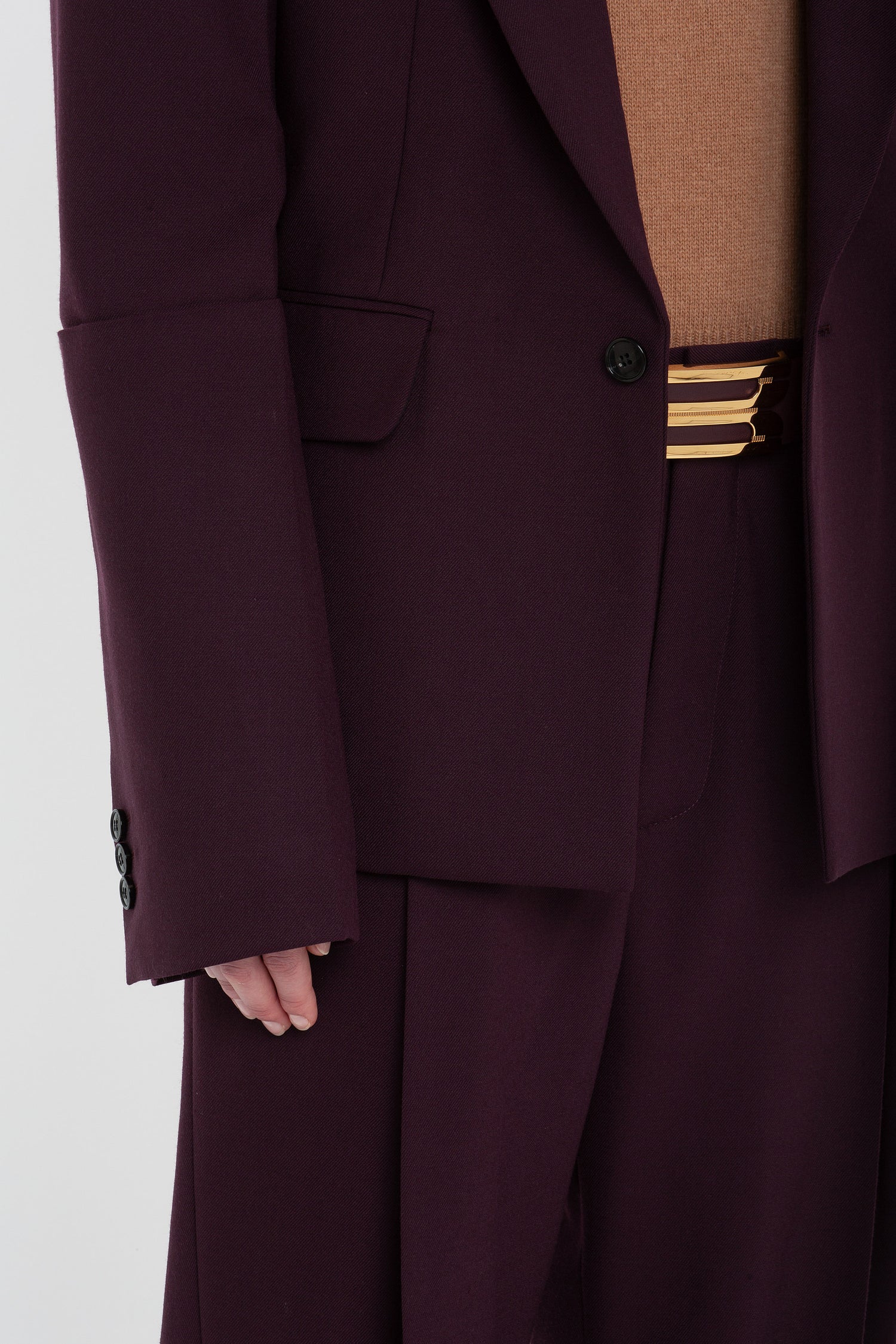 Close-up of a person wearing a dark purple, single-breasted blazer and matching pants with a peach-colored shirt. The Sleeve Detail Patch Pocket Jacket In Deep Mahogany by Victoria Beckham, made from recycled wool, has long sleeves, and there is a gold-colored belt buckle visible.