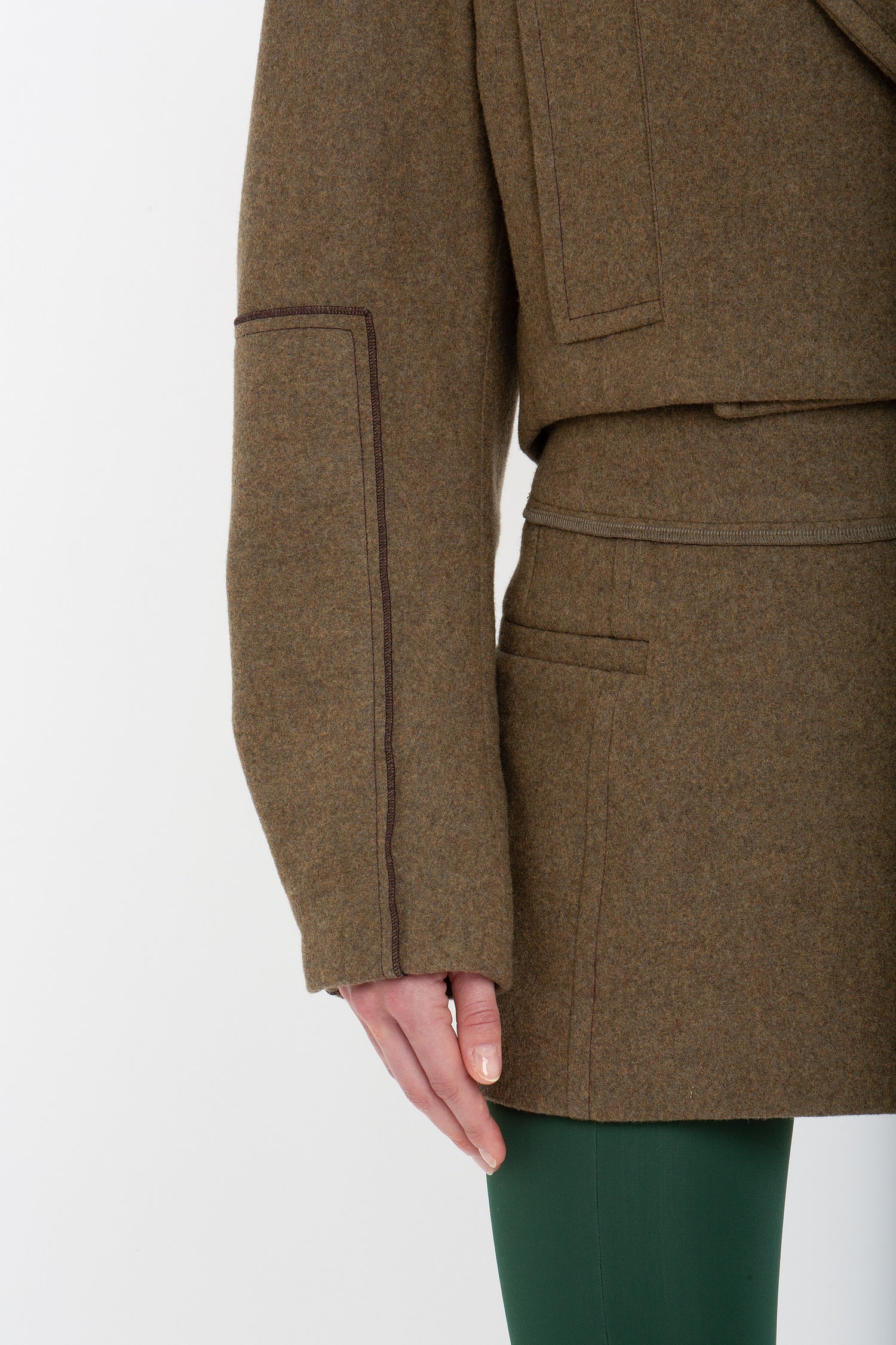 Close-up of a person wearing a brown coat of merino wool and a Victoria Beckham Tailored Mini Skirt In Khaki. Only the lower half of the coat and the upper part of the skirt are visible along with one hand touching the coat's pocket.