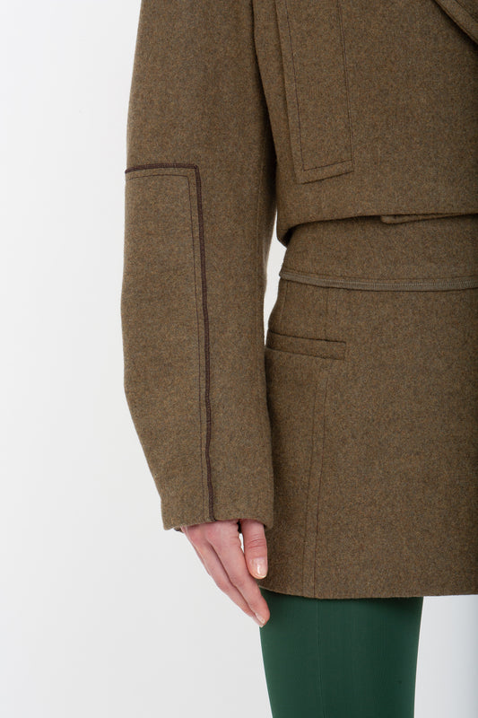 Close-up of a person wearing a brown coat of merino wool and a Victoria Beckham Tailored Mini Skirt In Khaki. Only the lower half of the coat and the upper part of the skirt are visible along with one hand touching the coat's pocket.