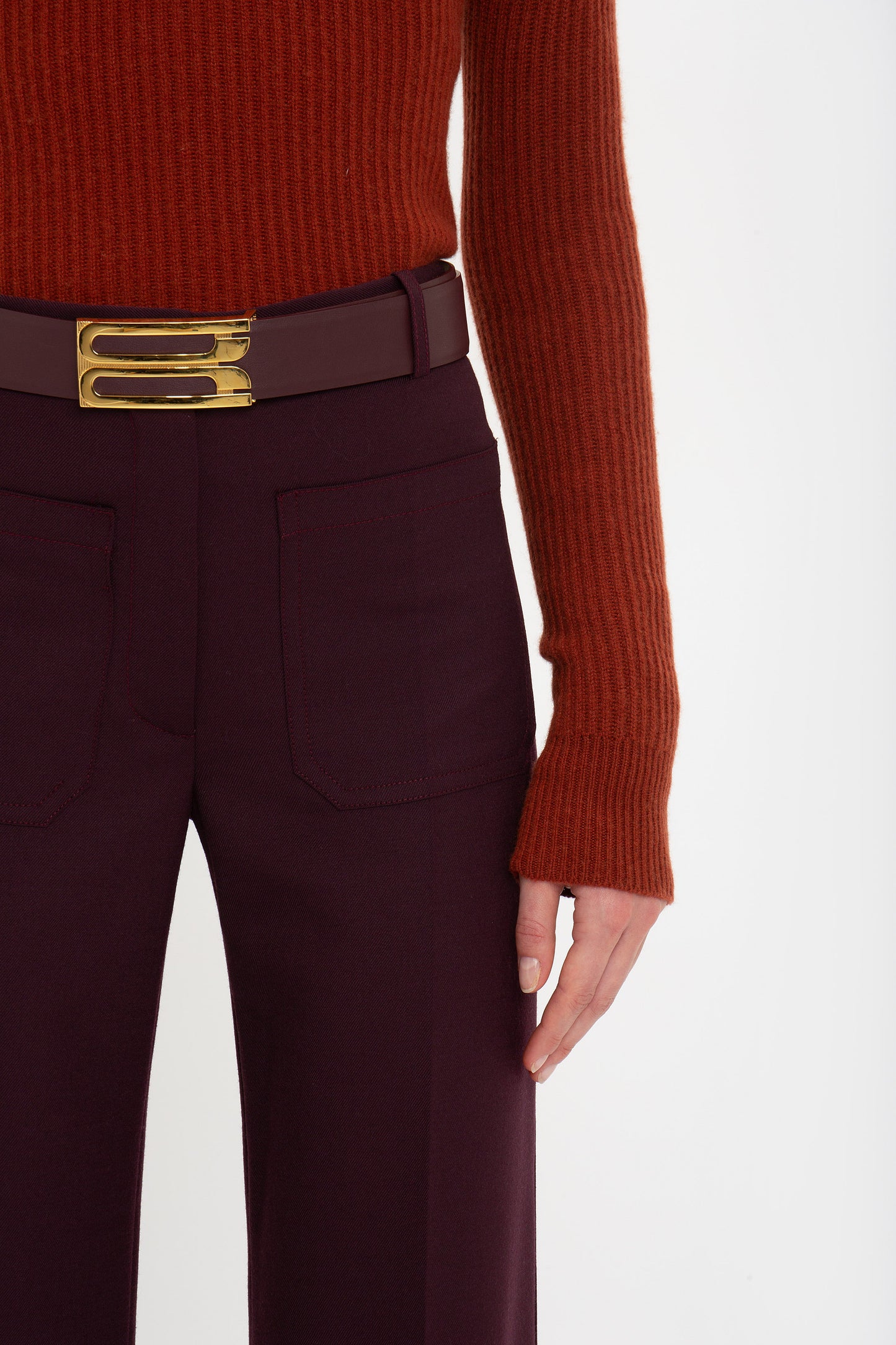 A person wearing a rust-colored ribbed sweater made of recycled wool, deep mahogany Victoria Beckham Alina Trouser In Deep Mahogany, and a burgundy belt with a gold buckle stands against a plain background. The right arm is slightly bent.