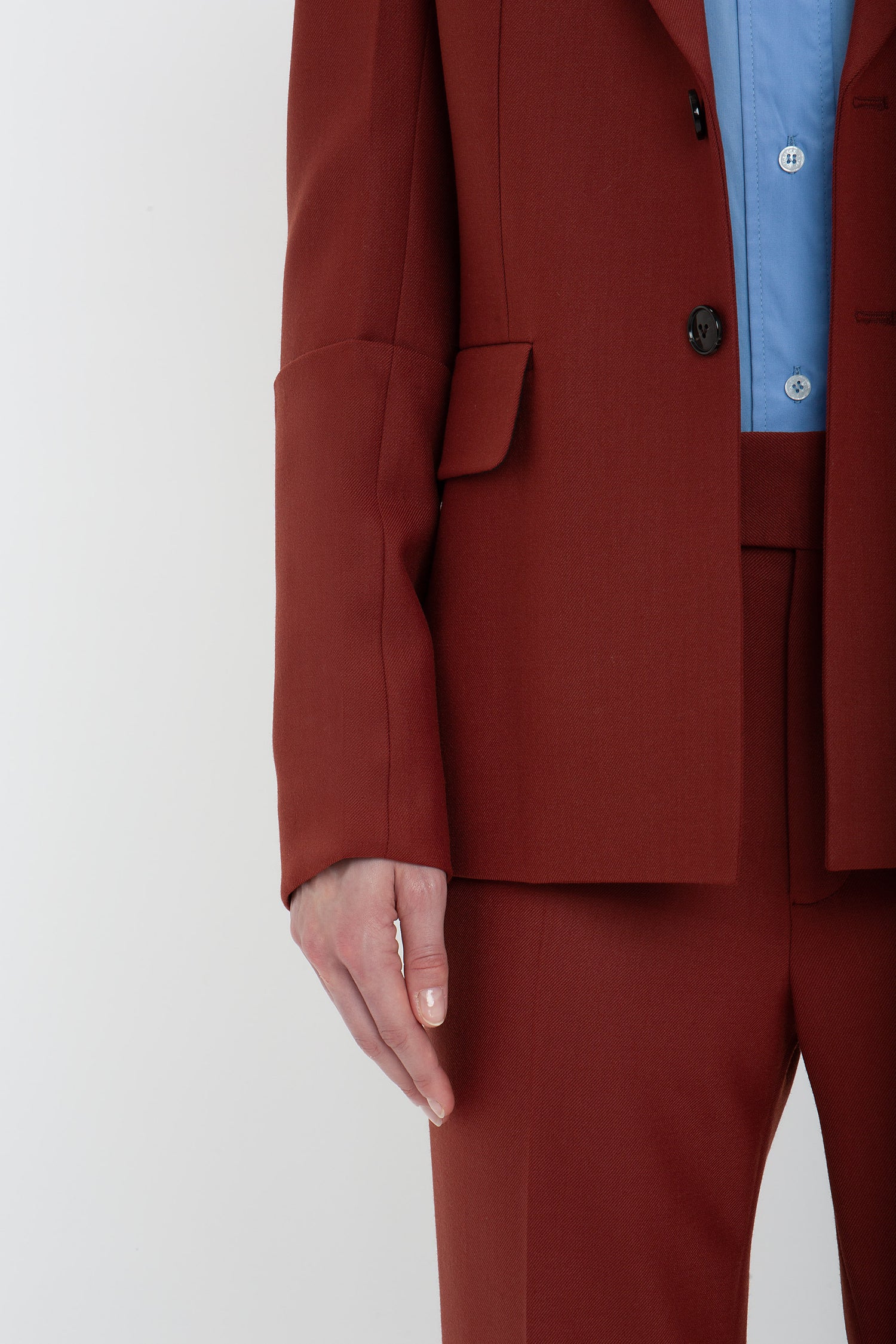 Close-up of a person wearing a red Sleeve Detail Patch Pocket Jacket In Russet by Victoria Beckham and pants with a light blue shirt. Their left hand is visible by their side, showcasing the contemporary detailing. The background is plain white.