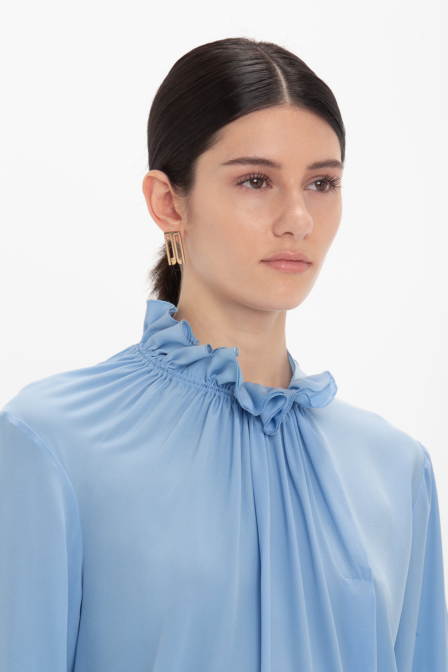 A person with long dark hair tied back wearing a light blue, crepe de chine Exclusive Ruffle Neck Blouse In Cornflower Blue by Victoria Beckham and gold earrings shaped like rectangles. The background is plain white.