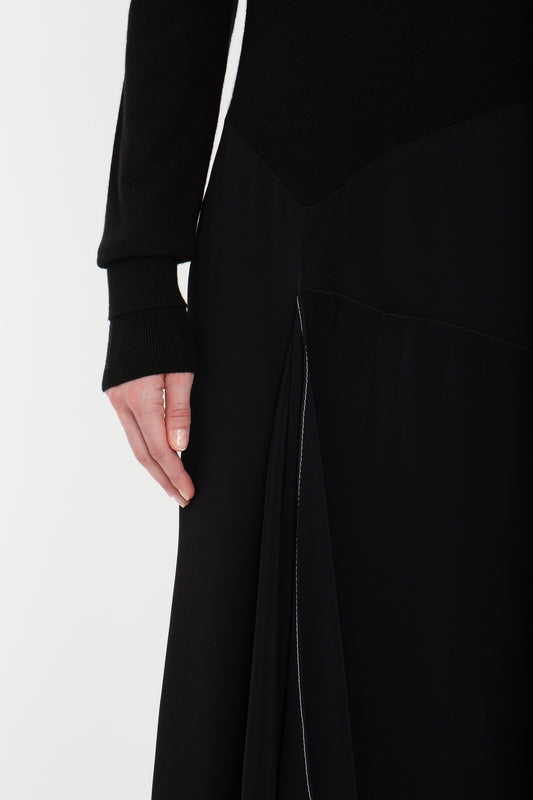 Close-up of a person wearing a black Henley Shirt Dress In Black by Victoria Beckham. One hand is visible, resting along the side. The dress features a subtle pleat detail and an asymmetric waist. The background is plain white.