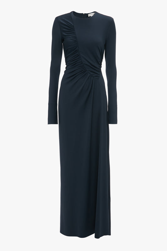 A long-sleeve, floor-length navy blue dress with a ruched detail on one side and a round neckline, reminiscent of the Victoria Beckham Ruched Detail Floor-Length Gown In Midnight.