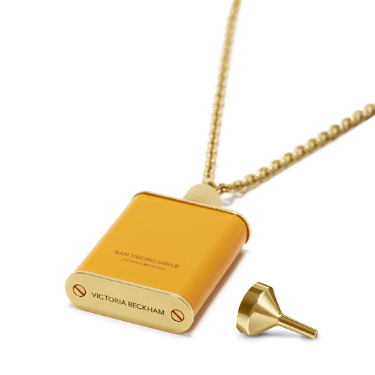 A gold flask-shaped pendant with "Victoria Beckham" inscribed, featuring a chain and a small funnel placed beside it, perfect for holding your favorite scent. This Perfume Bottle Necklace In San Ysidro Drive exemplifies gold brushed brass jewelry at its finest.