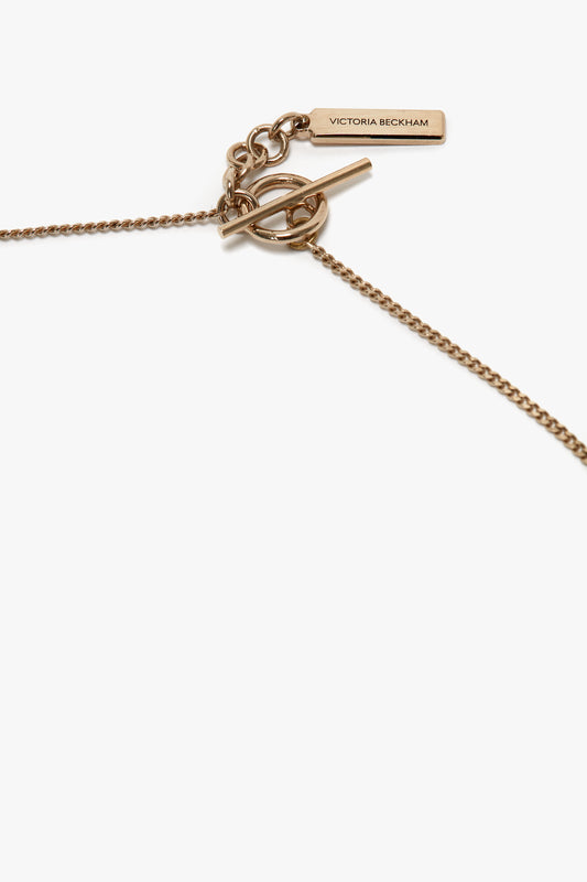 Close-up of an Exclusive Body Charm Necklace In Light Gold, featuring a toggle clasp and a small rectangular tag engraved with "Victoria Beckham." Crafted from 100% brass.