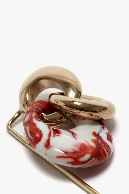 Close-up of the Exclusive Resin Charm Brooch In Light Gold-Orange by Victoria Beckham with intertwined metal hoops and a resin piece featuring red and white marbling, made in Italy.