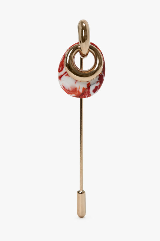 A light gold stick pin showcases a circular red and white marbled design, enhanced with a looped gold accent at the top. This elegant Victoria Beckham Exclusive Resin Charm Brooch In Light Gold-Orange is made in Italy.