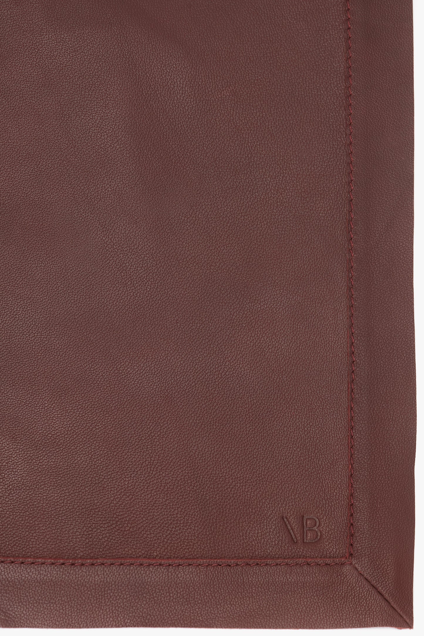 Close-up shot of a maroon leather square with detailed stitching along the edges, resembling the sophisticated Leather Scarf In Bordeaux by Victoria Beckham, and a small embossed logo in the bottom right corner.