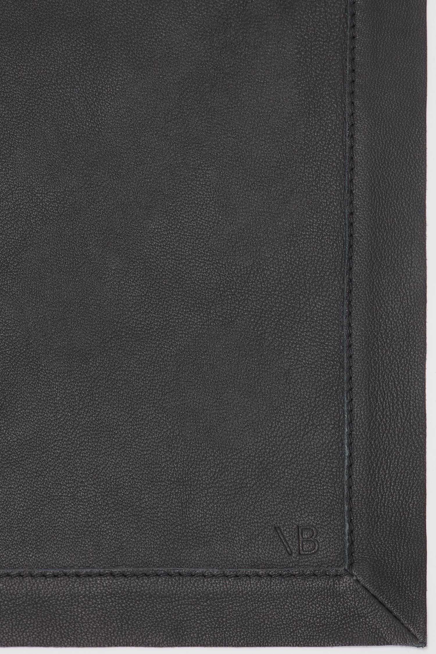 Close-up of a dark, textured leather scarf with visible stitching and the initials "VB" embossed at the bottom right corner, showcasing its classic black hue and supple finish. The product is the Victoria Beckham Leather Scarf In Black.