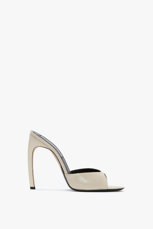 Side view of a beige luxury calf leather high-heeled mule with an open back and a seductive curved heel, set against a white background. The **Classic Mule In Macadamia Calf Leather by Victoria Beckham** takes center stage with its elegant design and premium material.