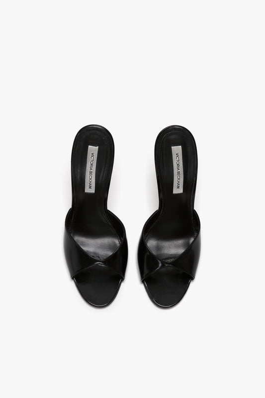 A pair of black, open-toe, slip-on women’s shoes with a crisscross design and seductive curved heel, insoles labeled "Classic Mule In Black Calf Leather by Victoria Beckham.
