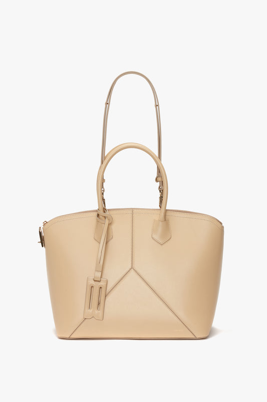 The Victoria Beckham Victoria Bag In Sesame Leather is a beige leather handbag with two handles and a luggage tag attached to one of them. Featuring leather panels, this stylish bag has a slightly structured shape, zip closure, and an adjustable strap for added convenience.