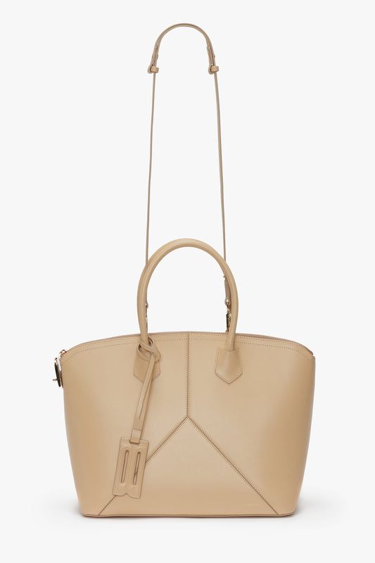 The Victoria Beckham Victoria Bag In Sesame Leather, featuring beige leather panels, boasts a shoulder strap, two handles, and a luggage tag accessory, displayed against a white background.