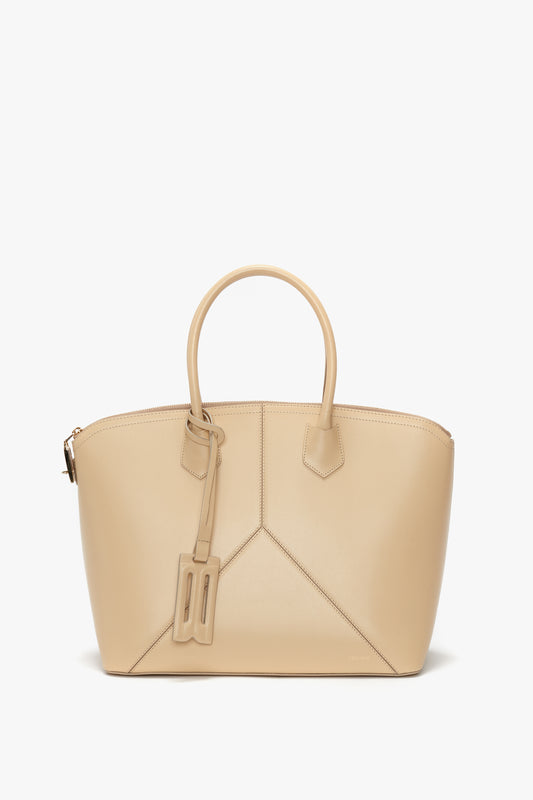 The Victoria Beckham Victoria Bag In Sesame Leather features a beige tote design with two handles, a luggage tag, and leather panels beautifully accenting the geometric front design against a white background.