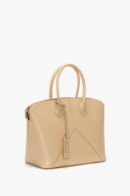 Introducing the Victoria Beckham Victoria Bag In Sesame Leather: a beige leather handbag with two handles and a geometric design crafted from elegant leather panels. It boasts a zipper closure at the top, a matching leather tag attached, and an option for an adjustable strap. All set against a pristine white background.