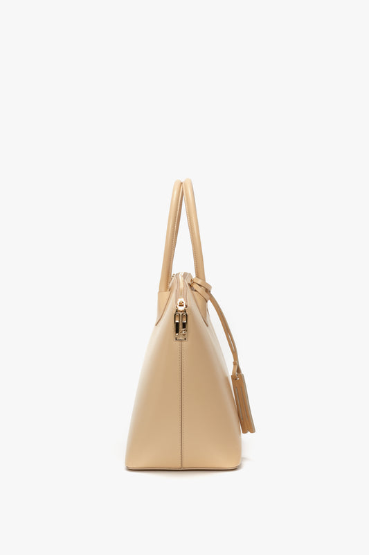 Side view of the Victoria Beckham Victoria Bag In Sesame Leather with a structured design and top handles. The bag features leather panels, a gold-tone zipper, a tassel detail on the side, and an adjustable strap for versatile styling.