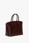 A burgundy leather Victoria Bag In Burgundy Leather by Victoria Beckham with two handles, gold hardware, a luggage tag, and an adjustable strap.