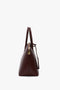 Side view of a closed dark brown Victoria Beckham Victoria Bag In Burgundy Leather with two handles, an adjustable strap, and a gold zipper on a white background.