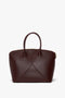 A Victoria Beckham Victoria Bag In Burgundy Leather with double handles, angular stitching on the front, an adjustable strap, and a zippered top closure.