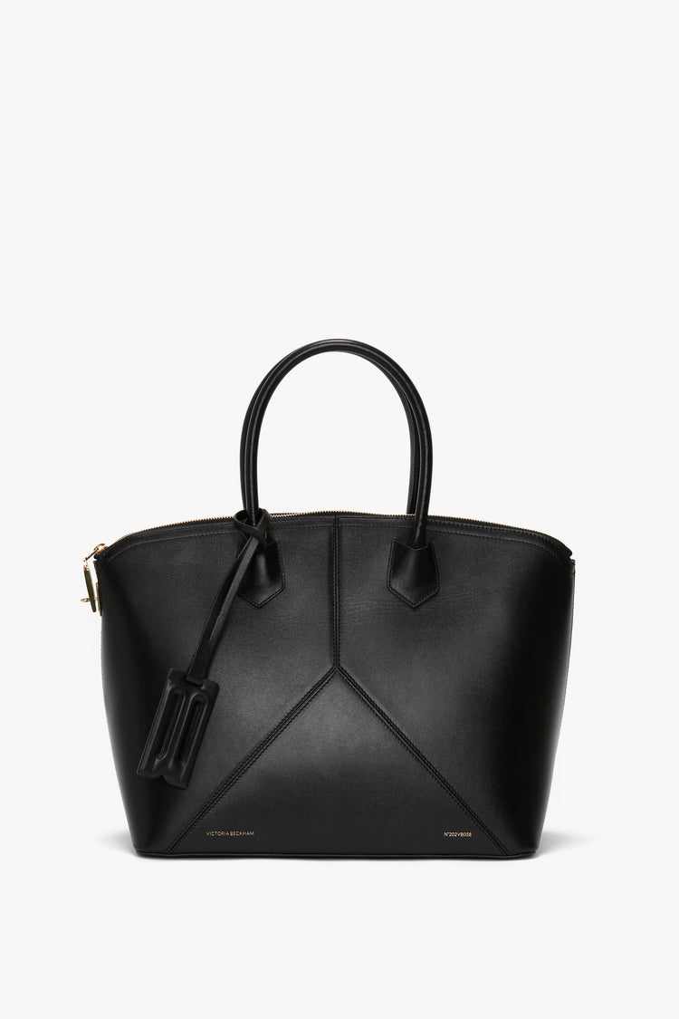 The Victoria Bag In Black Leather by Victoria Beckham is a versatile hold-all, crafted with black leather panels in a structured, trapezoidal design. It features handles, minimalistic details, and a small rectangular tag attached.