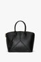 The Victoria Bag In Black Leather by Victoria Beckham is a versatile hold-all, crafted with black leather panels in a structured, trapezoidal design. It features handles, minimalistic details, and a small rectangular tag attached.