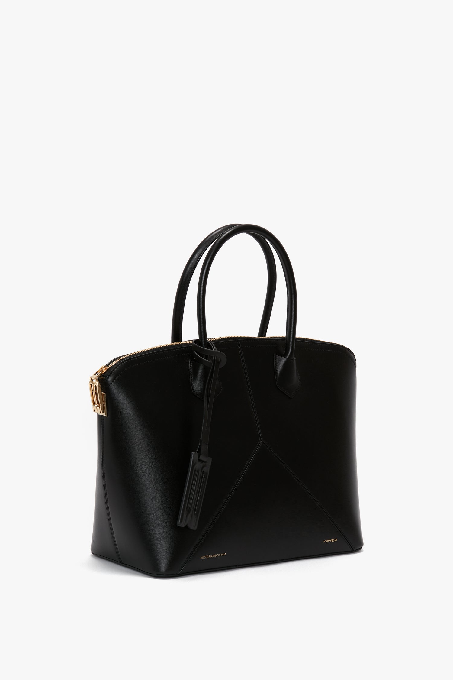 The versatile hold-all, the Victoria Bag In Black Leather by Victoria Beckham, features black leather panels, two handles, and a gold zipper. Its structured, angular design is complemented by a small luggage tag attached for added flair.