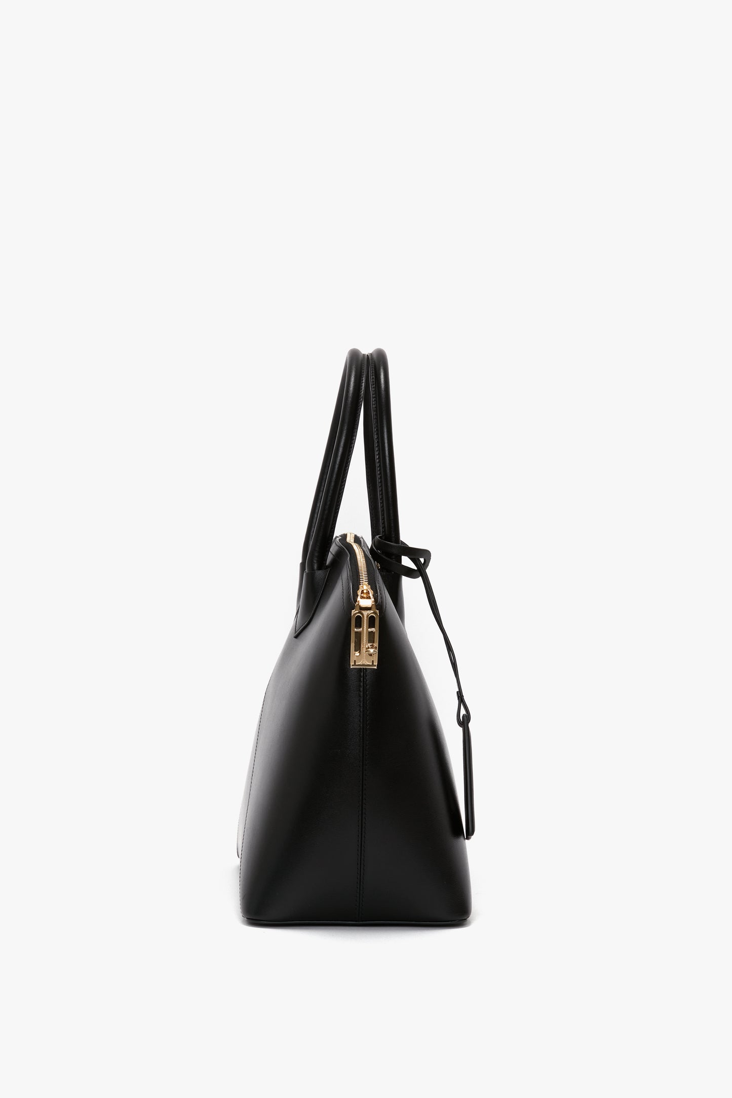 Side view of the Victoria Bag In Black Leather by Victoria Beckham, a versatile hold-all featuring black leather panels, gold zipper closure, and two handles against a white background.