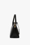 Side view of the Victoria Bag In Black Leather by Victoria Beckham, a versatile hold-all featuring black leather panels, gold zipper closure, and two handles against a white background.