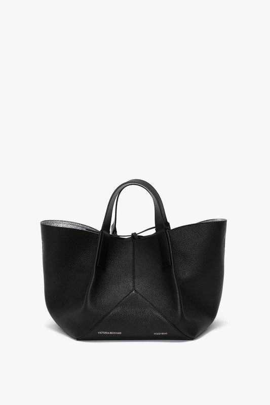 A black leather tote bag with luxurious sophistication, featuring two handles and a wide, open top. The W11 Mini Tote In Black Leather by Victoria Beckham boasts subtle brand names at the base.