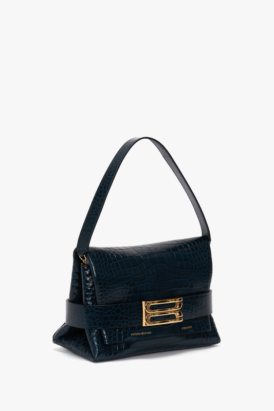 A B Pouch Bag In Croc Effect Midnight Blue Leather by Victoria Beckham, featuring a short handle, prominent gold clasp, and angular design. This handbag also includes a detachable shoulder strap for versatile styling.