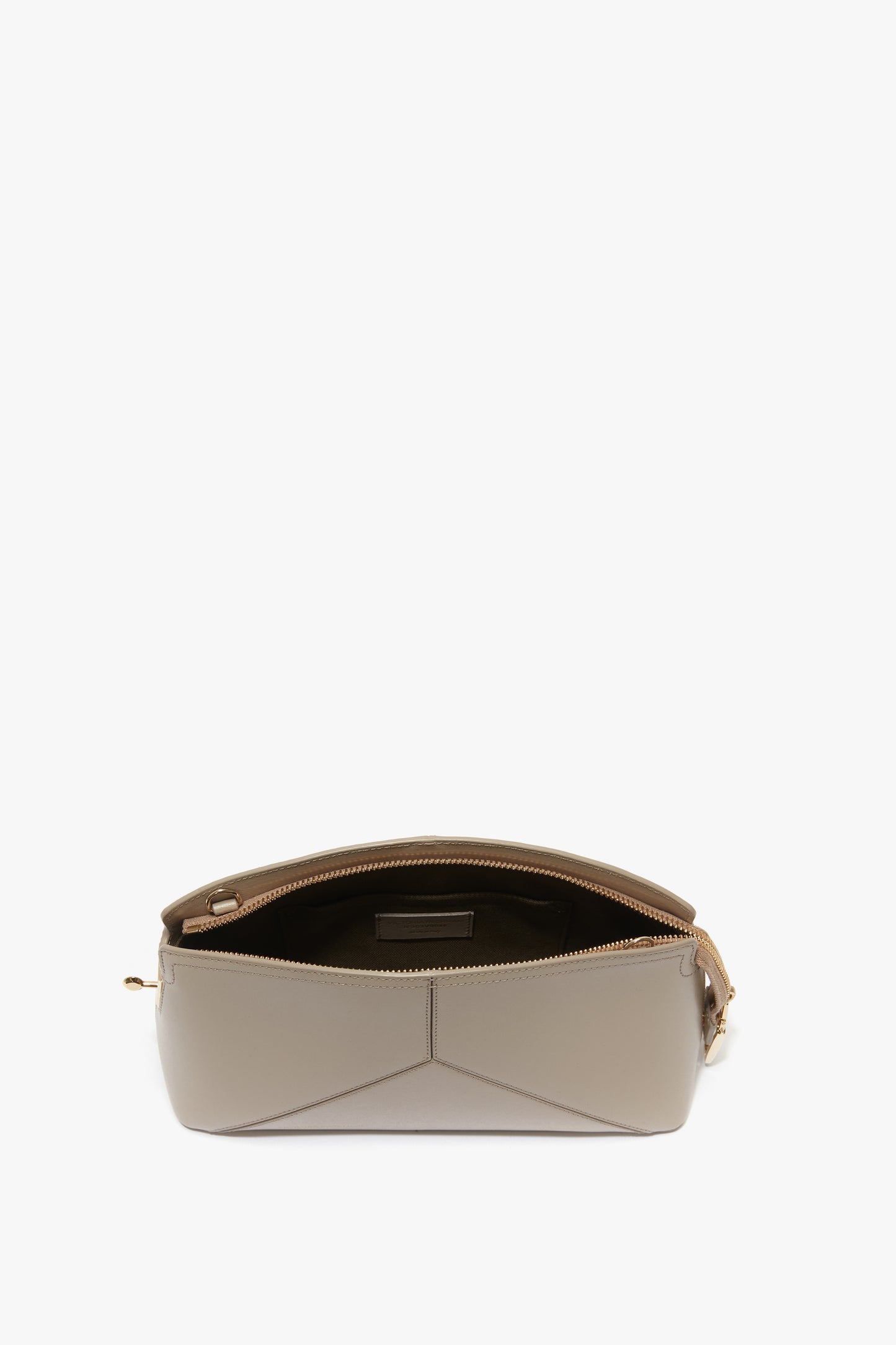 A Victoria Crossbody Bag In Taupe Leather is shown with its top zipper open, revealing an empty interior. The bag, from Victoria Beckham, features angular stitching on the front panel and a curved base for a structured design.