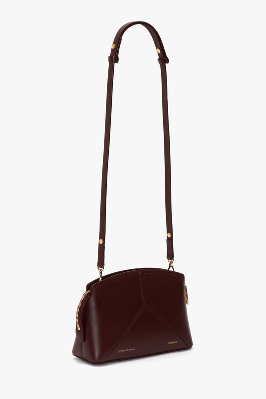 The Victoria Beckham Victoria Crossbody In Burgundy Leather is a structured calf leather bag featuring a long adjustable strap, gold-tone hardware, and a curved zipper closure on the top.