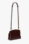 The Victoria Beckham Victoria Crossbody Bag In Burgundy Leather is a dark brown, structured calf leather bag featuring an adjustable strap, gold zippers, and a secure padlock closure.