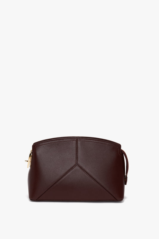 A small, dark burgundy leather handbag with a geometric design and a gold zipper. The bag has a structured calf leather finish, minimalist style, and a wrist strap on one side, perfect for those who appreciate the elegance of the Victoria Beckham Victoria Crossbody In Burgundy Leather.