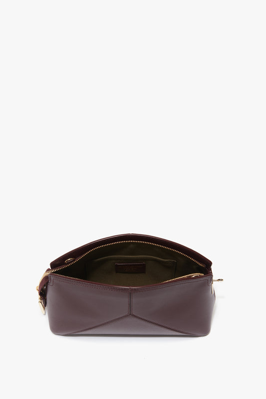 A small, burgundy leather purse with a central zip opening reveals an olive green interior lining. The Victoria Beckham Victoria Crossbody In Burgundy Leather has a structured shape with clean, geometric lines and a gold buckle on one side.
