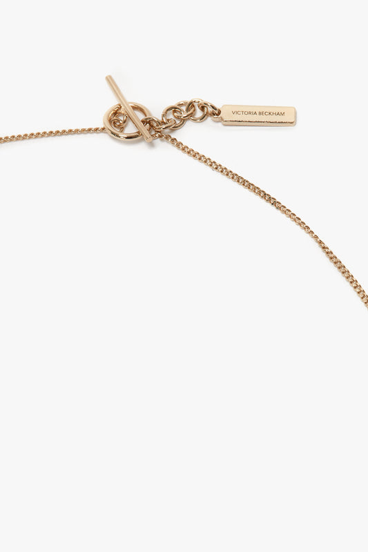 A 31cm-long, light gold-colored necklace featuring a minimalist chain and a toggle clasp with a rectangular tag inscribed "Exclusive Resin Pendant Necklace In Light Gold-White by Victoria Beckham".