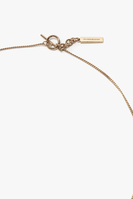An Exclusive Resin Pendant Necklace In Light Gold-Black featuring a small rectangular tag engraved with "Victoria Beckham" and a simple clasp mechanism, crafted from 100% brass, on a white background.