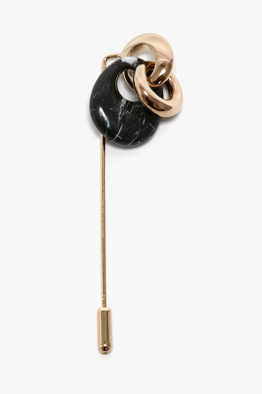 A Victoria Beckham Exclusive Resin Charm Brooch In Light Gold-Black featuring two light gold interlocking rings and a black teardrop-shaped stone on one end, with a long, straight 100% brass metal pin extending from the other end.