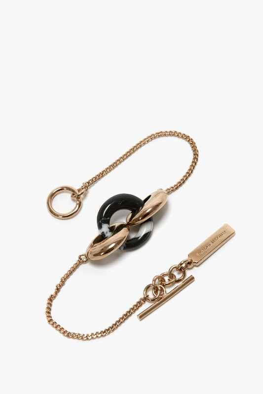 Victoria Beckham's Exclusive Resin Charm Bracelet In Light Gold-Black with a circular clasp, featuring a black and white looped detail and rectangular tag, crafted from 100% brass for added durability. This elegant piece can be styled as a resin charm bracelet for an extra touch of sophistication.