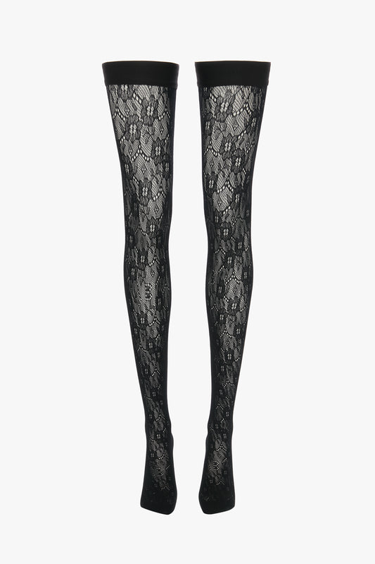 A pair of Exclusive VB Monogram Lace Hold-Ups In Black by Victoria Beckham, these black lace patterned thigh-high stockings feature a sheer finish and a solid black band at the top, displayed vertically.