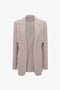 A light beige or grey women's blazer with a structured design, featuring sharp lapels, a single breast pocket, and two front pockets. This menswear-inspired tailored jacket has a sleek fit and appears to be made of smooth fabric. The Double Panel Front Jacket In Rose Quartz by Victoria Beckham.