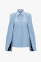 A light blue long-sleeve blouse with a collar and layered, draping sleeves featuring black accents near the cuffs. The Victoria Beckham Pleat Detail Raglan Shirt In Oxford Blue contemporary silhouette boasts pleat details and a concealed button-front closure for a sleek finish.