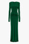 Victoria Beckham's Circle Detail Open Back Gown In Emerald is a green long-sleeve gown crafted from jersey fabric with shoulder pads and a gathered design at the waist. The dress is floor-length with an exposed back and a fitted silhouette.