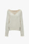 A Victoria Beckham V-Neck Jumper In Natural, a cream-colored, long-sleeve cable-knit sweater with a contrasting tan color accent at the neckline. Crafted from soft lambswool, the jumper features a relaxed fit with ribbed cuffs and hem.