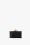 A Victoria Beckham Frame Flower Minaudiere Bag in Black with a gold flower clasp and a detachable shoulder strap lies elegantly against a plain white background.