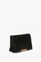 A Victoria Beckham Puffy Jumbo Chain Pouch In Black Leather with a folded design, embellished with a small gold geometric pattern on the lower front edge, displayed against a plain white background.