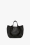 A black grained leather tote bag with two short handles, a structured bottom, and subtle gold branding near the base. This W11 Medium Tote Bag In Black Leather by Victoria Beckham, inspired by Victoria Beckham's timeless style, combines luxury with everyday practicality.