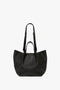 A black, grained leather W11 Medium Tote Bag In Black Leather with short handles and an additional adjustable longer strap. The bag features a textured surface and gold Victoria Beckham logo embossing at the bottom.